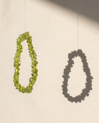 ⁠Suspended zero-metal raw green peridot crystal bracelet with sharp shadow play on a minimalist wall, mindful energy jewelry by STILLMANTLE⁠
