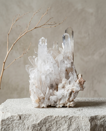 Raw clear quartz crystal cluster with natural matrix resting on a rough stone pedestal next to a dry branch, minimalist zen meditation room decor by STILLMANTLE⁠