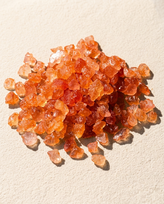 Macro view of raw orange sunstone unpolished chips with subtle geological glitter on light sand, minimalist quiet luxury grounding mineral parcel by STILLMANTLE