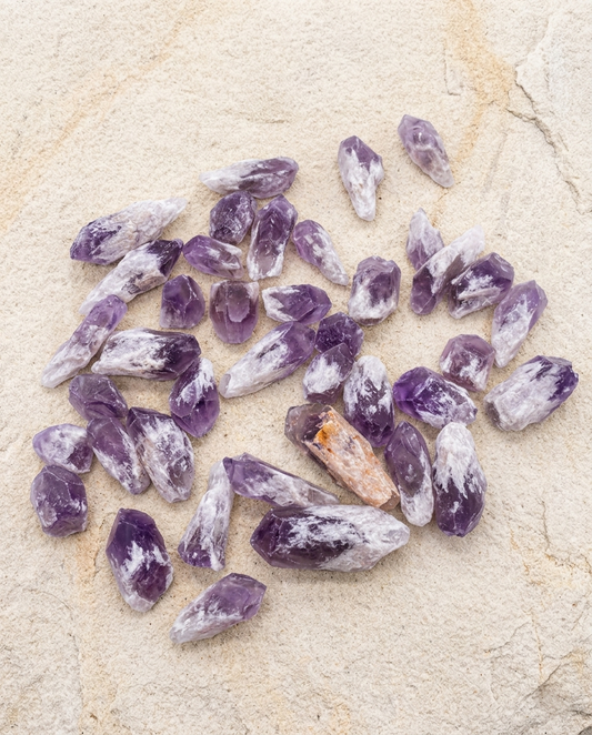 Collection of raw amethyst root points showing natural color zoning and unpolished crystal terminations on light textured sandstone, wabi-sabi unhewn healing mineral parcel by STILLMANTLE
