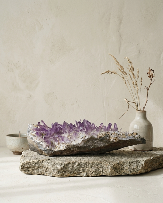 Massive horizontal Vera Cruz amethyst specimen on weathered stone base, styled with a minimalist ceramic vase and dried botanicals, wabi-sabi earth element statement sculpture by STILLMANTLE⁠