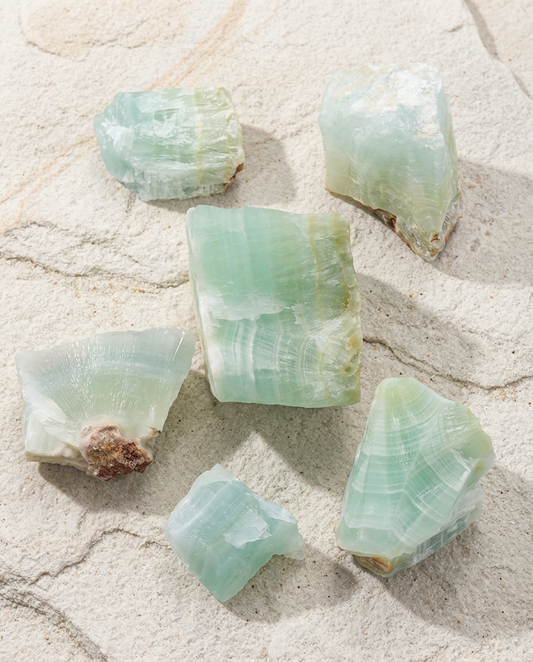Raw Caribbean Calcite stone chunks featuring natural layered bands of light sea-foam green and sandy white on a textured light stone surface. A curated earth mined gemstone bundle containing raw orbicular agate fragments.