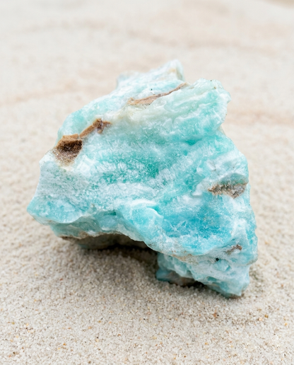 A rugged, natural electric blue Hemimorphite rough stone with crystalline textures and minor brown matrix patches, sitting on a bed of fine pale sand.