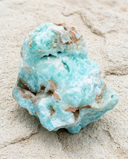A bright Caribbean blue Hemimorphite raw crystal displaying natural botryoidal formations and small brown matrix details, placed against a textured sandy background.