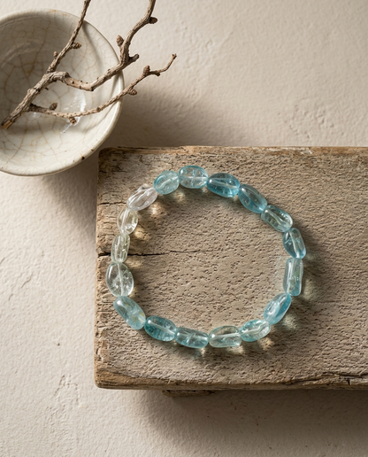 Close-up photography of a natural gradient Beryl gemstone bracelet on a clean, off-white textured background. The asymmetrical tumbled stones show a seamless ombré transition from clear transparent Goshenite to icy blue Aquamarine, revealing authentic internal mineral inclusions.⁠