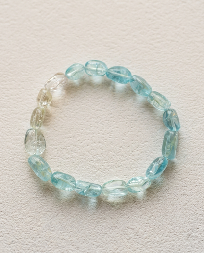 Natural gradient Aquamarine and Goshenite beryl bracelet resting on a weathered, textured wooden block. Minimalist wabi-sabi curatorial staging with a cracked ceramic raku bowl and dried twigs, emphasizing the raw, glacial-blue crystal clarity.⁠