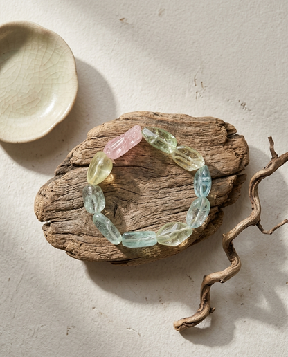Curated collection of raw pastel crystal fragments arranged as a bracelet on reclaimed wood, paired with a ceramic bowl, minimalist organic modern jewelry aesthetic.