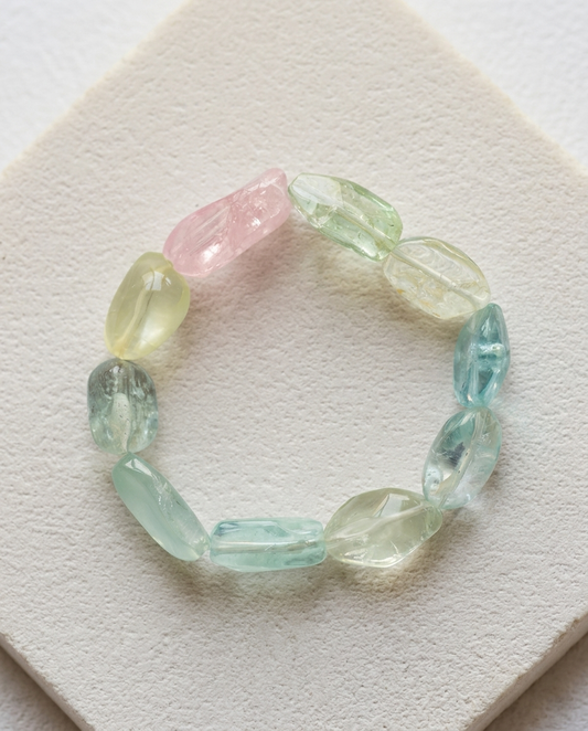 Close-up of a raw tumbled pastel mineral bracelet with varied gemstone shapes, displayed on a clean textured surface, high-end organic crystal jewelry, minimalist fashion accessory.