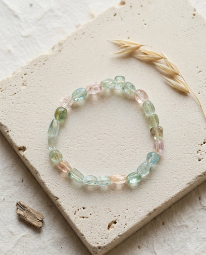 ⁠High clarity multi-color beryl bracelet featuring pink morganite and blue aquamarine, resting on a square travertine tile with dried oats and driftwood, luxury mineral jewelry.⁠
