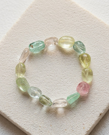 Macro close-up of a high transparency pastel mixed beryl bracelet resting on a geometric textured stone plinth, unheated organic tumbled crystals.⁠