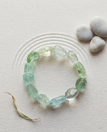 Natural cool-toned beryl gemstone bracelet with aquamarine and green beryl, minimalist styled on raked zen sand with smooth grey pebbles and a dry leaf, organic modern jewelry aesthetic.⁠