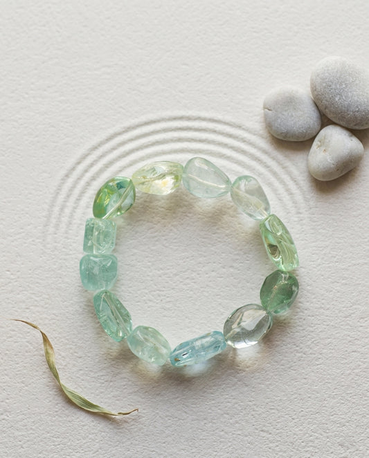 Natural cool-toned beryl gemstone bracelet with aquamarine and green beryl, minimalist styled on raked zen sand with smooth grey pebbles and a dry leaf, organic modern jewelry aesthetic.⁠