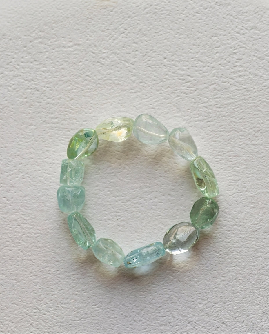 Macro close-up of a high transparency mixed cool-toned beryl bracelet resting on a textured white surface, unheated organic tumbled aquamarine crystals.
