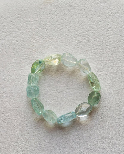 Macro close-up of a high transparency mixed cool-toned beryl bracelet resting on a textured white surface, unheated organic tumbled aquamarine crystals.
